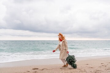 Fototapeta premium Redhead woman Christmas tree sea. Christmas portrait of a happy redhead woman walking along the beach and holding a Christmas tree in her hands. Dressed in a light coat, white suit and red mittens.