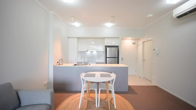 Small apartment dinging and kitchen area interior. Grey carpet, grey lounge room sofa. Metal stainless steel fridge. split system aircon
