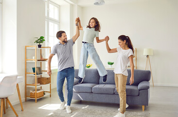 Happy family of three having fun together. Cheerful parents and child spending time at home. Joyful mother and father holding daughter's hands while she jumps off sofa in living room - obrazy, fototapety, plakaty