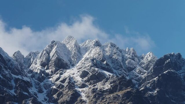 Timelapse of Rugged Snowy Mountains with beautiful clouds rolling over with a blue sky. It's steep and rocky on a windy day. The Remarkables, New Zealand.