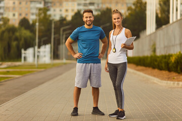 Portrait of a happy sporty couple in sportswear with stopwatch standing after sport jogging exercises in city park and looking cheerful at camera. Outdoors training and fitness in nature concept.