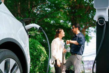 Young couple recharge electric car's battery from charging station in outdoor green city park in springtime. Rechargeable EV car for sustainable environmental friendly urban travel lifestyle.Expedient