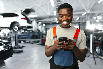 Young African mechanic man wearing uniform using mobile phone in a car service