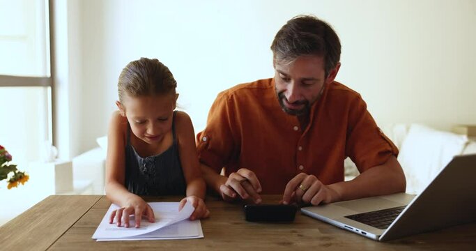 Little loving daughter help to father with work, sit together at table managing family budget and finances, calculate expenses and incomes paying household bills using e-banking system, check savings