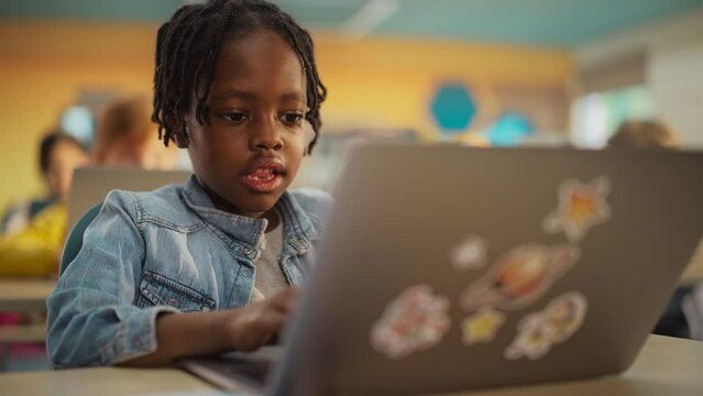 Smart Little Black Boy with Dreadlocks Sitting Behind a Desk with a Laptop Computer in Primary School. Young Attentive African Man Writing Down Notes, Working on an Online Exercise