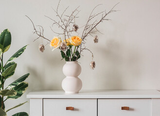Easter bouquet of yellow roses and dry twigs with glass Easter eggs on a white wooden chest of...