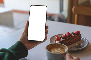 Mockup image of a woman holding mobile phone with blank screen while drinking coffee and eating dessert in cafe