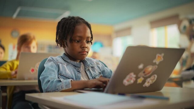 Portrait of a Cute Little African Boy with Stylish Hair Sitting Behind a Desk in Class in Elementary School. Young Pupil Using a Laptop Computer while Listening to a Teacher with Other Kids