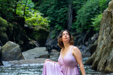 A young woman in beautiful pink dress rests on the banks of the river and rainforest.