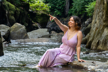 A young woman in beautiful pink dress rests on the banks of the river and rainforest.