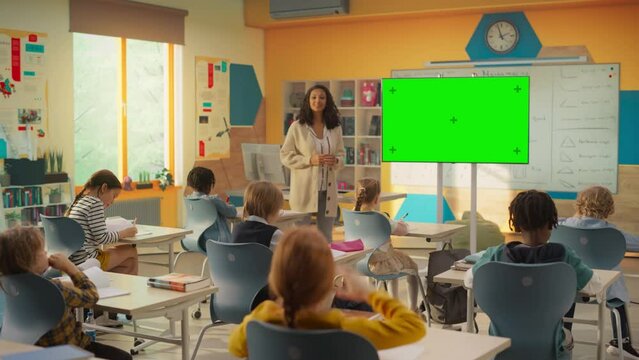 Caring Primary School Teacher Using a TV with Green Screen Display, Explaining an Exercise to a Classroom Full of Bright Diverse Children. Little Girl Raising Her Hand and Asking Tutor a Question