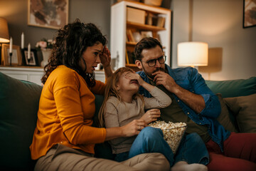 Parents and daughter immersed in a film, creating cherished memories with a bowl of popcorn - obrazy, fototapety, plakaty