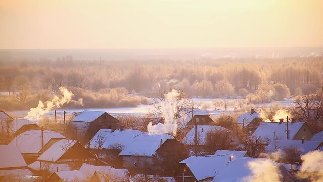 Smoke and steam from chimneys on the roofs of houses in the village with stove heating. Frosty winter morning against the backdrop of the sun and beautiful nature. Copy space for text, countryside