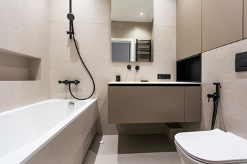 Bathroom in a modern style with gray and white tiles. There is a large mirror with luminous lamps, tabletop with wooden drawers and sink, bath with shower and glass partition, towel rack and a hanger.