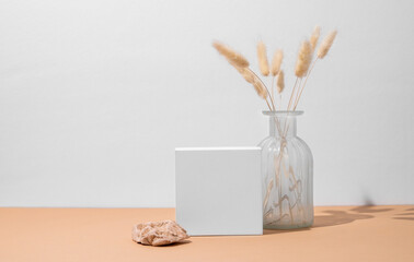 White empty box and vase with dried flowers on a beige background with a hard shadow.