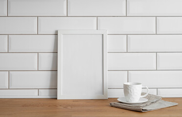 Fototapeta na wymiar A frame with a blank canvas against a white tile wall and on a wooden tabletop with cup of tea.