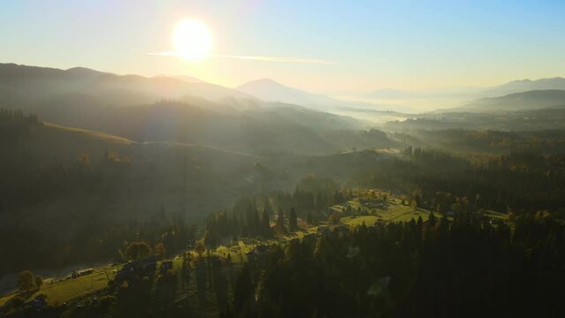 Wooded hills of Carpathian mountains in Ukraine with small traditional village houses at sunset. Brightly illuminated with setting sun evergreen woods with scattered local homes at fall season