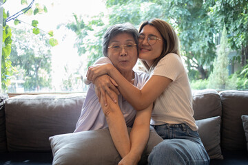 Happy asian Thai elder mother and daughter sitting on couch and cuddles, smiling laughing and enjoy moment together.