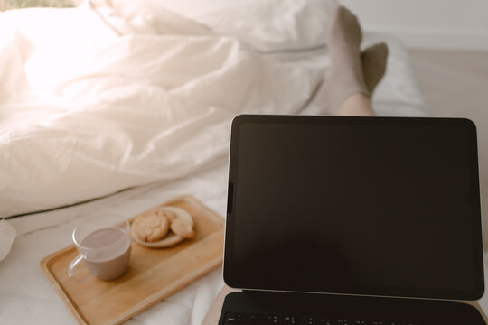Image of laptop showing black screen display putting on lap, woman using tablet and working while eating cookie and coffee on white bed at day time.