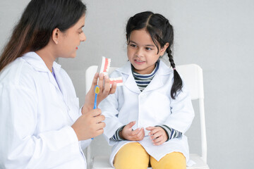 Cute Asian little girl in doctor coat learning how to brush her teeth and gums on a jaw model from young Indian dentist with toothbrush at dentist's clinic. Oral and Dental Health