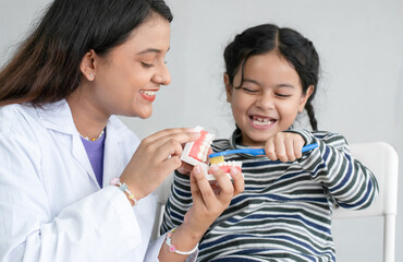 Cute Asian little girl learning how to brush her teeth and gums on a jaw model from young Indian dentist with toothbrush at clinic. Oral and Dental Health. Selective focus on dentist