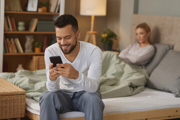 Man with smartphone in bedroom, upset woman in the background