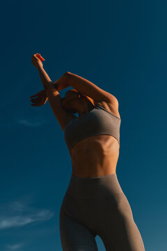Slender woman in gray yoga suit poses against blue sky.