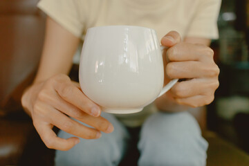 Close up of woman two hands holding white coffee cup, showing milk tea glass while sitting on brown sofa couch at home.