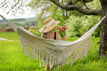 beautiful woman with sun hat, lying in a hammock on a meadow, holding a bunch of tulips in her hands, summer, spring, shot in nature - Powered by Adobe