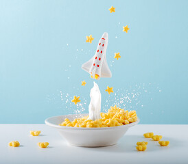 Small space rocket takes off from breakfast plate with cornflakes and milk on pastel blue...