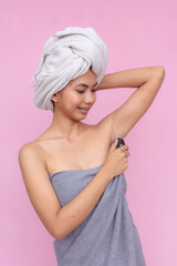 A cheerful young Asian woman in a towel applies roll-on antiperspirant to her armpits against a vibrant pink background.