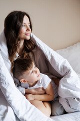 Toddler boy in pajama sitting near young mother under blanket on bed at home