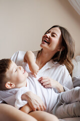 Cheerful woman in shirt hugging toddler son on bed in morning