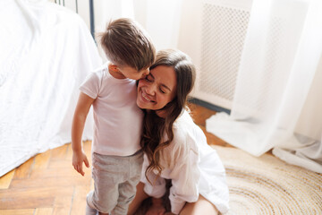 Toddler boy in pajama kissing cheerful mother in bedroom in morning