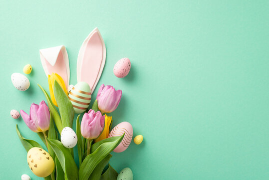 Easter happiness in photo! Overhead perspective of a bouquet adorned with tulips and vibrant eggs, complemented by quirky bunny ears. Displayed on mint-green background, ready for personalized message