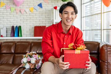 Young asian man with gift red box in his hands and bouquet, sitting on the sofa in his room on...