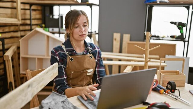 Attractive young blonde woman carpenter engrossed in a lively online video call amidst rustic carpentry workshop, surrounded by timber and woodwork tools.