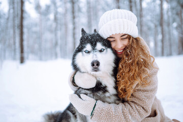 A young woman in warm clothes walking her dog in a picturesque snowy forest. Woman laughing and playing with pet in the park. Domestic dog concept.