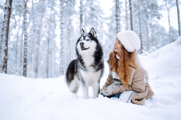 A young woman in warm clothes walking her dog in a picturesque snowy forest. Woman laughing and playing with pet in the park. Domestic dog concept.
