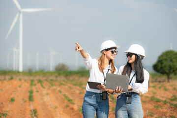 Two asian engineer women holding laptop and working in wind turbine farm with blue sky background