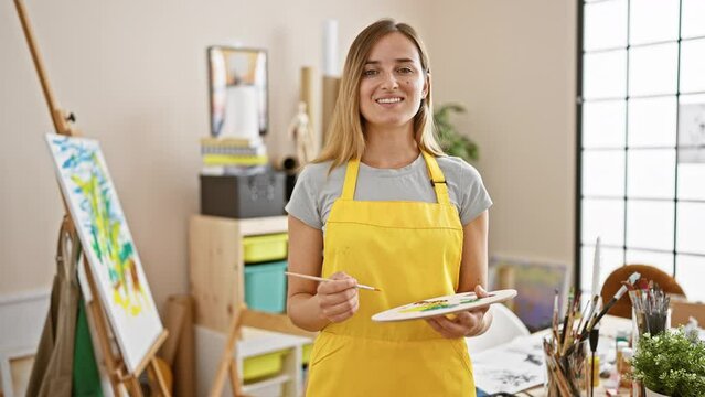 Attractive young blonde woman artist confidently smiling, holding a paintbrush and palette in the lively art studio environment