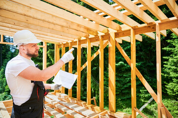 Surveyor inspecting construction plans of wooden two-story building being erected. Bearded man in...