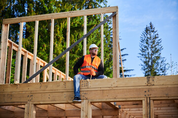 Carpenter constructing wooden frame two-story house near the forest. Bearded man holding hammer, dressed in protective helmet and orange safety vest. Concept of ecological modern construction.