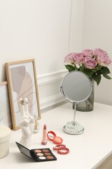 Mirror, cosmetic products, perfume and vase with pink roses on white dressing table in makeup room