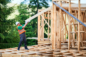 Carpenter constructing wooden-framed house near the woods. Man holding large truss, wearing work clothes and helmet. Concept of modern and eco-friendly building.