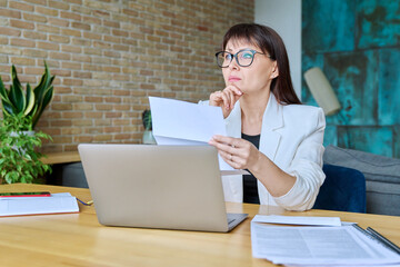 Serious mature businesswoman reading official letter, sitting in office