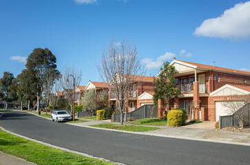 A suburban road lined with two-story residential townhouses on small blocks of land in an Australian neighbourhood. A road in a residential suburb with brick homes.