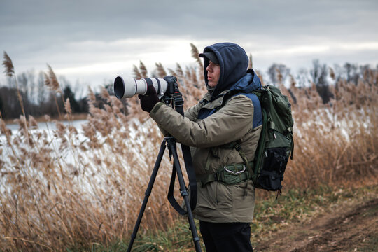Photographer is setting up camera on tripod outdoors. Man photographing landscape or wildlife at lake in winter
