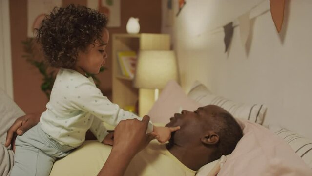 Medium side shot of African American father lying in bed and asking for kisses from little Biracial toddler girl who kissing him on mouth and cheeks in bedroom in eveningMedium side shot of African Am