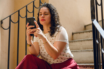 young attentive latin woman reading message on phone sitting outside on stairs at home - obrazy, fototapety, plakaty